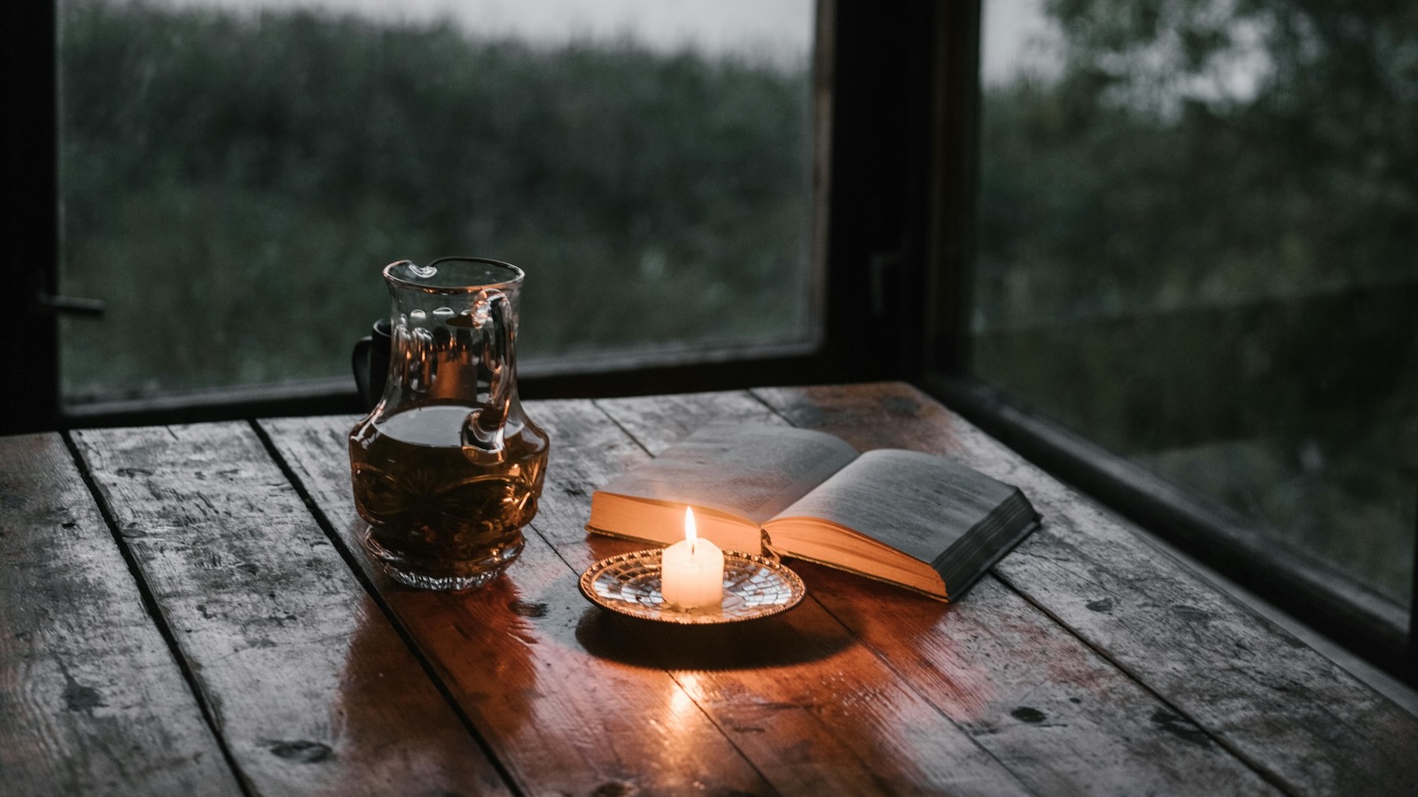 white candle book and decanter on rustic wooden table near window overlooking a lake at dusk featured image for wmarkwhitlock.com promoting Beloved Strangers Devotions and Prayers for Estranged Christian Parents